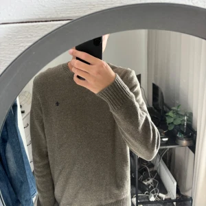 Olivgrön stickad tröja från Morris - Säljer en snygg olivgrön stickad tröja från Morris i storlek M. Tröjan har rund hals, ribbade muddar och ett diskret Morris-emblem broderat på bröstet. Perfekt för dig som gillar stilrena och klassiska plagg med skön passform.