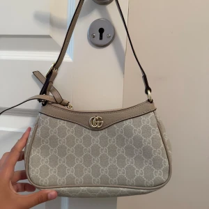 Begagnad väska  - Snygg beige axelväska  med klassiskt  och guldfärgade logodetalj framtill. Väskan har justerbar axelrem och dragkedja upptill. Materialet är en mix av skinn och canvas