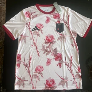 Japan blommig fotbollströja Adidas XL - Snygg Japan fotbollströja från Adidas i vit med röda blommor och blad över hela tröjan. Kortärmad med röd krage och ärmslut, badge med Japan-flagga och JFA-logga på bröstet. Tillverkad i lätt, tekniskt material som andas.