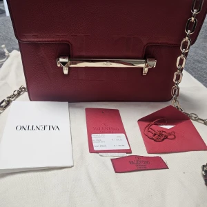 Röd axelväska Valentino Garavani - Snygg röd axelväska från Valentino Garavani i skinn med präglad logga på framsidan. Väskan har en kedja i silverfärgad metall och ett stilrent metallspänne med nitar. Perfekt storlek för det viktigaste och har exklusiv känsla.