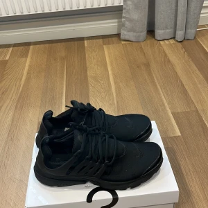 Svarta Nike Presto sneakers - Nike Presto sneakers i helsvart färg med snygg, strömlinjeformad design. Ovandel i mjuk syntet och mesh, med platt sula och rund tå. Skorna har snörning och en cool plastdetalj på sidorna för extra support. Perfekt för dig som gillar stilrena och sportiga sneakers.