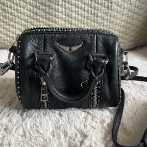Svart handväska från Zadig  - Cool svart handväska från Zadig Voltaire i skinn med silverfärgade nitar längs kanterna och detaljerade dragkedjor framtill. Väskan har både handtag och axelrem.