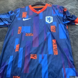 Hollands landslagströja blå/orange Nike - Säljer en officiell Holland fotbollströja från Nike i blått med orange detaljer och mönster. Tröjan har korta ärmar, KNVB-emblem på bröstet och vit Nike-logga. Unik grafisk design med inslag av rött och ljust blått. Perfekt för fans av Nederländernas landslag.