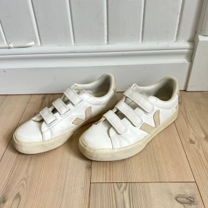 Vita sneakers från Veja - Säljer ett par klassiska vita sneakers från Veja med beige detaljer och kardborreband💕