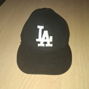 Svart LA keps med broderad logga - Svart keps med vit broderad LA-logga framtill och texten 'Los Angeles' baktill. Justerbart spänne i metall bak. Klassisk böjd skärm och normal passform. Perfekt för dig som gillar sportig stil.