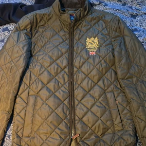 Olivgrön quiltad jacka från Morris - Snygg olivgrön quiltad jacka från Morris med broderad logga och brittisk flagga på bröstet. Jackan har dragkedja framtill, ståkrage och två snedställda fickor. Perfekt för kyliga dagar.