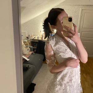 Studentklänning  - Säljer en superfin vit kortklänning med spets och blommiga broderier. Klänningen har v-ringning, halvtransparenta ärmar och fina volangdetaljer. Perfekt för sommarens tillfällen eller studentfirande. Figurnära passform och vackra spetskanter.