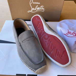 Grå loafers från Christian Louboutin - Snygga grå loafers från Christian Louboutin med ovandel i mocka och klassisk röd sula. Skorna har dekorativ söm runt tån och flätad detalj längs sulkanten. Perfekta för dig som vill ha en stilren och exklusiv look.