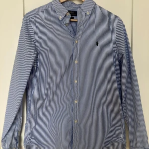 Blårandig skjorta från Polo Ralph Lauren - Klassisk blårandig skjorta från Polo Ralph Lauren i slim fit-modell. Skjortan har långa ärmar, knappar framtill och en broderad logga. Skulle säga att den sitter som en small. Använd ett fåtal gånger, otroligt bra skick utan några fläckar eller skador.