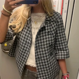 Grå och svart rutig kappa med trekvartsärm - Säljer en snygg grå och svart rutig kappa med stora knappar och trekvartsärmar. Jackan har en klassisk krage och är i en kortare modell som ger en trendig look. Perfekt för dig som vill sticka ut med ett tidlöst mönster.