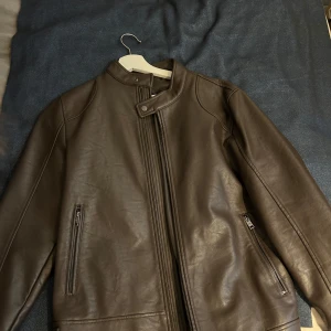 Brun skinnjacka från Mango Man - Säljer en brun skinnjacka från Mango Man i klassisk modell med ståkrage och dragkedja. Jackan har två snedställda fickor med dragkedja och en knapp vid halsen. Perfekt för dig som gillar stilrena och tidlösa plagg. Säljer pga att den inte kommit till användning 