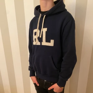 Mörkblå hoodie från Ralph Lauren - Säljer en snygg mörkblå hoodie från Ralph Lauren med stor RL-logga på framsidan. Tröjan har en klassisk känguruficka och långa ärmar med ribbade muddar. Perfekt för en avslappnad stil.