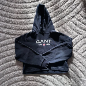 Mörkblå hoodie från GANT - Säljer en mörkblå hoodie från GANT i barnstorlek. Tröjan har huva och långa ärmar, samt ribbade muddar vid ärmslut och nederkant. Perfekt för en avslappnad stil. 