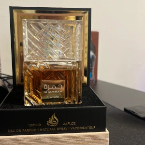 Khamrah Eau de Parfum från Lattafa - Khamrah från Lattafa är en lyxig parfym i en elegant fyrkantig glasflaska med guldiga detaljer och ett geometriskt mönster på locket. Flaskan rymmer 100 ml och kommer i en svart och guldig förpackning som utstrålar exklusivitet.