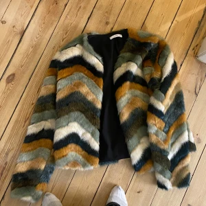 Flerfärgad pälsjacka från Mango - Säljer en snygg pälsjacka från Mango med zigzag-mönster i grönt, svart, vitt, beige och orange. Jackan har lång ärm och är fodrad med svart tyg på insidan.