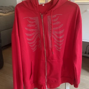 Röd hoodie med strassdetaljer - Säljer en röd hoodie med dragkedja och huva. Framsidan är dekorerad med strass i ett coolt mönster. Tröjan har fickor framtill och justerbar dragsko vid huvan. Perfekt för dig som vill sticka ut lite extra!
