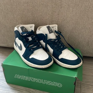 Nike Air Jordan 1 Mid blå och vita sneakers - Säljer ett par Nike Air Jordan 1 Mid sneakers i blått och vitt. Skorna har klassisk snörning, rund tå och vit sula. Perfekta för dig som gillar streetwear och vill ha en ikonisk look. Boxen medföljer.