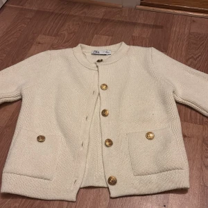 Krämvit stickad cardigan jacka från Zara - Säljer en krämvit stickad cardigan jacka från Zara med guldknappar framtill och på fickorna. Jackan har rund halsringning och långa ärmar. Perfekt för lager-på-lager och enkel att styla.