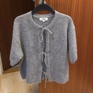 Grå stickad kofta med knyt fram från gina tricot - Supermjuk grå stickad kofta från Gina Tricot med korta ärmar och tre knytband framtill. Perfekt att slänga över en topp. Rund halsringning och avslappnad passform.