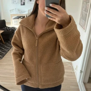 Beige teddyjacka med dragkedja - Mysig beige teddyjacka med dragkedja framtill och hög krage. Passformen är normal och materialet är mjukt och fluffigt.