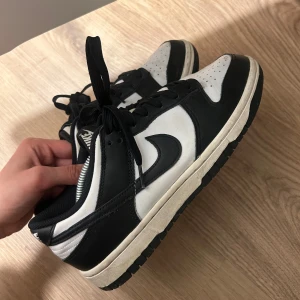 Nike Dunk Low svartvita sneakers - Säljer ett par klassiska Nike Dunk Low sneakers i svart och vitt. Skorna har svart snörning, vit tåbox och sula samt svart Nike-logga på sidan. Perfekta för dig som gillar streetwear och vill ha en stilren look.