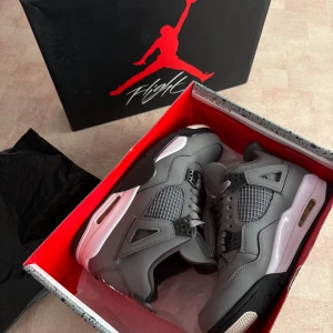 Gråa Nike Air Jordans 4 Retro - Säljer ett par gråa Nike Air Jordans 4 Retro med svarta och vita detaljer. Skorna har klassisk snörning, meshpaneler och Jumpman-logga på plösen och hälen. Snygg och sportig design med platt sula och rund tå.
