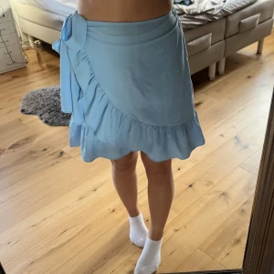 Ljusblå omlottkjol från Vero Moda - Supersöt ljusblå omlottkjol från Vero Moda med volangkant och knyt i midjan. Kjolen har en kort längd och är perfekt för varma dagar. Tillverkad i ett lätt och luftigt material. På framsidan finns några små knottror, vilket inte är något man tänke på med kjolen på🫶🏼🫶🏼