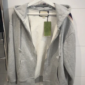 Grå hoodie med dragkedja från Gucci - Säljer en grå hoodie från Gucci ! HELT NY !med dragkedja och huva. Jackan har fickor framtill och en vit, röd och blå rand med Gucci-logga längs ärmen. Perfekt för en avslappnad och trendig stil. ! PRIS KAN DISKUTERAS ! STORLEK M !
