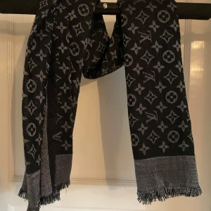 Svart och grå halsduk från Louis Vuitton - Snygg svart och grå halsduk från Louis Vuitton med ikoniskt monogrammönster. Halsduken har fransar i ändarna och är tillverkad i en lyxig blandning av silke och ull. Perfekt accessoar för att lyfta din outfit.