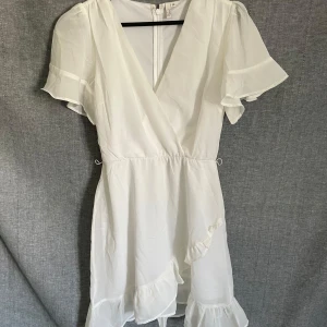 Vit omlottklänning med volang - Superfin vit omlottklänning med volangdetaljer och korta ärmar. Klänningen har v-ringning och lätt, luftig känsla – perfekt för varma dagar eller speciella tillfällen. Stängs med dragkedja i ryggen.