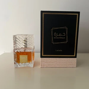 Khamrah Eau de Parfum från Lattafa - Khamrah från Lattafa är en parfym i en elegant, kristalliknande glasflaska med bärnstensfärgad vätska. Förpackningen är svart med guldiga detaljer och en beige botten. Parfymen är Eau de Parfum. Cirka 85-90ml kvar