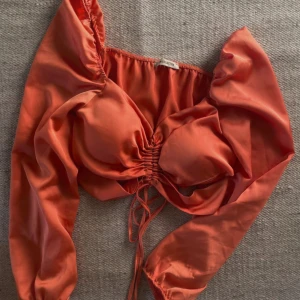 Korallfärgad satinblus - Korallfärgad blus i satin med långa ärmar och knytning framtill. Toppen har en snygg v-ringning och rynkade detaljer vid bysten för en feminin look.