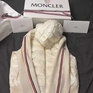 Vit pufferjacka med stickade detaljer från Moncler - Pris går att förhandla!!                             Exklusiv vit pufferjacka från Moncler med stickade ärmar och sidor. Jackan har huva, dragkedja och klassiska Moncler-ränder i rött, vitt och blått längs ärmarna. Broderad Moncler-logga på ärmen. Perfekt för dig som vill ha en stilren och sportig look.