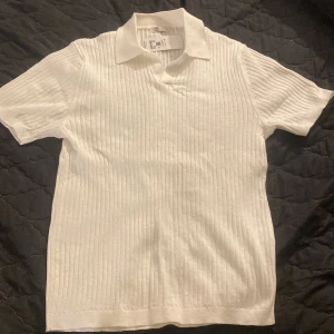 Vit stickad pikétröja - Säljer en stilren vit stickad pikétröja med klassisk krage och korta ärmar. Tröjan har ett diskret ribbat mönster som ger en snygg struktur. Perfekt för en clean och fräsch look.