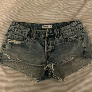 Blå jeansshorts med lite slitningar från Pull&Bear - Säljer ett par blå jeansshorts från Pull&Bear med slitna detaljer och fransig kant. Shortsen har klassisk femficksdesign, låg midja och knappar bak på fickorna. Perfekta för sommaren!