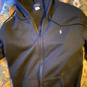 Svart hoodie med dragkedja  - Svart hoodie med dragkedja från Polo Ralph Lauren. Klassisk design med huva och vit broderad logga på bröstet. Perfekt för en avslappnad och stilren look.