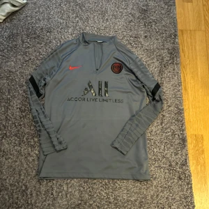 Grå långärmad PSG fotbollströja från Nike - Säljer en grå långärmad PSG fotbollströja från Nike med svarta detaljer och röd Nike-logga. Tröjan har klubbmärke på bröstet och tryck med 'Accor Live Limitless' samt 'Visit Rwanda' på ryggen. Perfekt för träning eller match. Den ena fickan har tappat sin dragkedja så går inte att öppna!