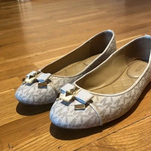 Vita ballerinaskor med rosett från Michael Kors - Säljer ett par vita ballerinaskor från Michael Kors med diskret MK-mönster, quiltad innersula och dekorativ rosett framtill med guldfärgade detaljer. Perfekta för en stilren och elegant look.