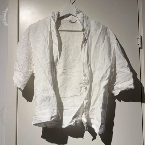 Vit linneskjorta med kort ärm - Säljer en luftig vit skjorta i linne med korta ärmar och klassisk krage. Skjortan har knappar framtill och en avslappnad passform, perfekt för varma dagar.