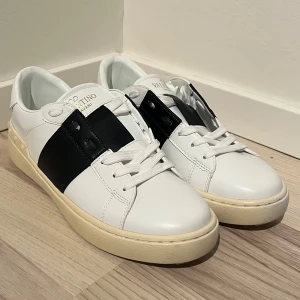 Vita Valentino Open sneakers med svart detalj - Säljer ett par snygga Valentino Open sneakers i vitt med bred svart rem över mitten. Skorna har vita snören och ikoniska nitar runt hälen. Perfekta för dig som gillar stilrena och exklusiva sneakers. Sparsamt använda.