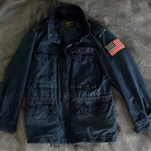 Polo Ralph Lauren field jacka - Säljer en mörkblå jacka från Ralph Lauren med unik denim still. Jackan har flera fickor framtill, axelklaffar och dragkedja med knappslå. Perfekt som vår/sommar jacka.