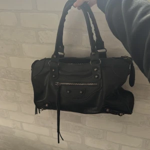 Svart handväska med nitar och dragkedja - Säljer en svart handväska med coola metalldetaljer och dragkedja framtill. Väskan har dubbla handtag och dekorativa spännen, perfekt för dig som gillar en edgy stil. Rymmer det mesta du behöver till vardagen.