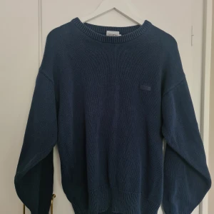 Mörkblå stickad tröja från Landy - Säljer en mörkblå stickad tröja från Landy. Tröjan har rund halsringning, breda ribbade muddar vid ärmslut och nederkant samt diskret logga på bröstet.