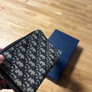 Dior axelväska med monogrammönster - Säljer en snygg axelväska från Dior med klassiskt monogrammönster i svart och grått. Väskan har justerbar axelrem, dubbla dragkedjor och flera fack inuti för kort och småsaker. Perfekt för dig som vill ha en stilren accessoar med lyxig känsla. Kvitto, låda,  påse och bäställningsorder finns 