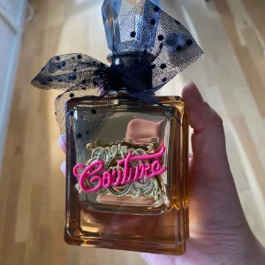 Couture parfymflaska med svart rosett - Säljer en lyxig parfym från Juicy Couture i en fyrkantig, bärnstensfärgad flaska med dekorativ svart prickig rosett och guldiga detaljer. Framsidan har en rosa Couture-logga i 3D. Perfekt för dig som gillar snygga och unika flaskor. Oanvänd