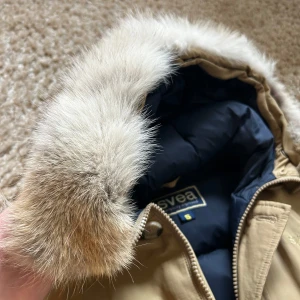 Beige parkajacka med pälskant från Svea - Säljer en beige parkajacka från Svea med avtagbar huva och pälskant. Jackan har dragkedja och knappar framtill, stora fickor och justerbar midja. Perfekt för kyliga dagar och riktigt mysig att bära.