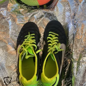 Svarta och neongröna fotbollsskor från Nike - Säljer ett par svarta fotbollsskor från Nike med neongröna detaljer och snörning. Skorna har rund tå och platt sula med fasta dobbar, perfekt för spel på gräsplan. Swoosh-logga på sidan och insida.