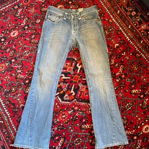 True Religion Joey Super T jeans - Klassiska blå jeans från True Religion, modell Joey Super T. Byxorna har kontrastsömmar, utsvängda ben och detaljerade bakfickor med knappar. Snygg vintage-look med skön fade och ikonisk etikett bak.