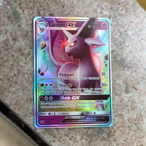 Pokémonkort Espeon GX - Säljer ett holografiskt Espeon GX Pokémonkort med 200 HP. Kortet har attacker som Psybeam, Psychic och Divide GX. Färgglad design med Espeon i fokus och glittrig yta. Perfekt för samlare eller Pokémon TCG-spelare.