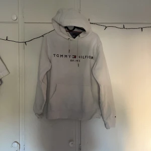 Vit hoodie från Tommy Hilfiger - Vit hoodie från Tommy Hilfiger med tryckt logga och text på bröstet. Klassisk huva med dragsko och känguruficka framtill. Perfekt för en avslappnad stil.